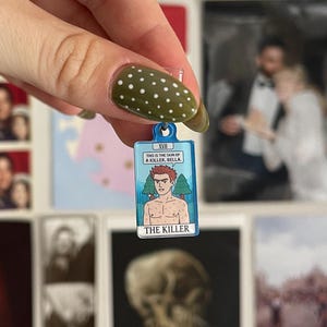 May include: A small rectangular enamel charm featuring a cartoon illustration of a shirtless person with red hair and green trees in the background. The text reads "THIS IS THE SKIN OF A KILLER, BELLA" and "THE KILLER" with the Roman numeral XVII at the top.