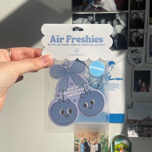 May include: Blueberry-shaped air freshener with the text "Air Freshies" and "you're doing great sweetie." The air freshener is light blue with a bow and two blueberries with smiling faces. The packaging says "for the car, home, office or wherever you are!"
