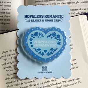 May include: A light blue phone grip and e-reader accessory with a heart-shaped design. The heart features the words "HOPELESS ROMANTIC" and floral accents. The accessory is on a light blue card with the Liv Lee Designs Co. logo.