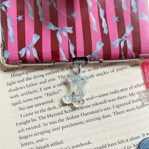 Op de afbeelding: Transparante telefoonhoes met een roze en rode streepjespatroon, versierd met lichtblauwe strikken en sterren. Een kleine, heldere bedel met een stripfiguur in een blauwe jas hangt aan de hoes. Een popsocket met een lichtblauw ontwerp is ook zichtbaar.
