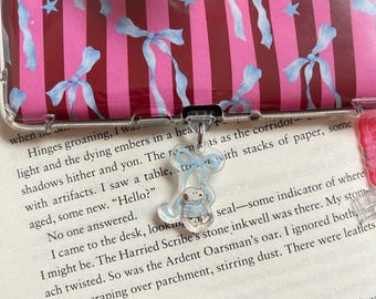 May include: Clear phone case with a pink and red striped pattern, adorned with light blue bows and stars. A small, clear charm with a cartoon character in a blue coat hangs from the case. A pop socket with a light blue design is also visible.