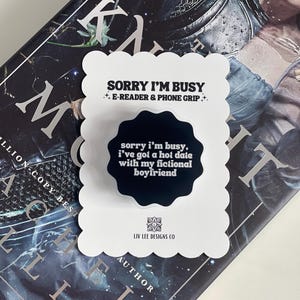Puede incluir: Un soporte para teléfono blanco con borde festoneado con el texto "SORRY I'M BUSY" y "E-READER & PHONE GRIP." Un elemento circular negro dice "sorry i'm busy, i've got a hot date with my fictional boyfriend." El soporte está sobre un libro.