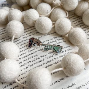 May include: Two enamel pins with detailed designs, one of a horse and the other of a mythical creature, rest on an open book. A garland of white felt balls surrounds the pins. The pins are colorful and the felt balls are soft.