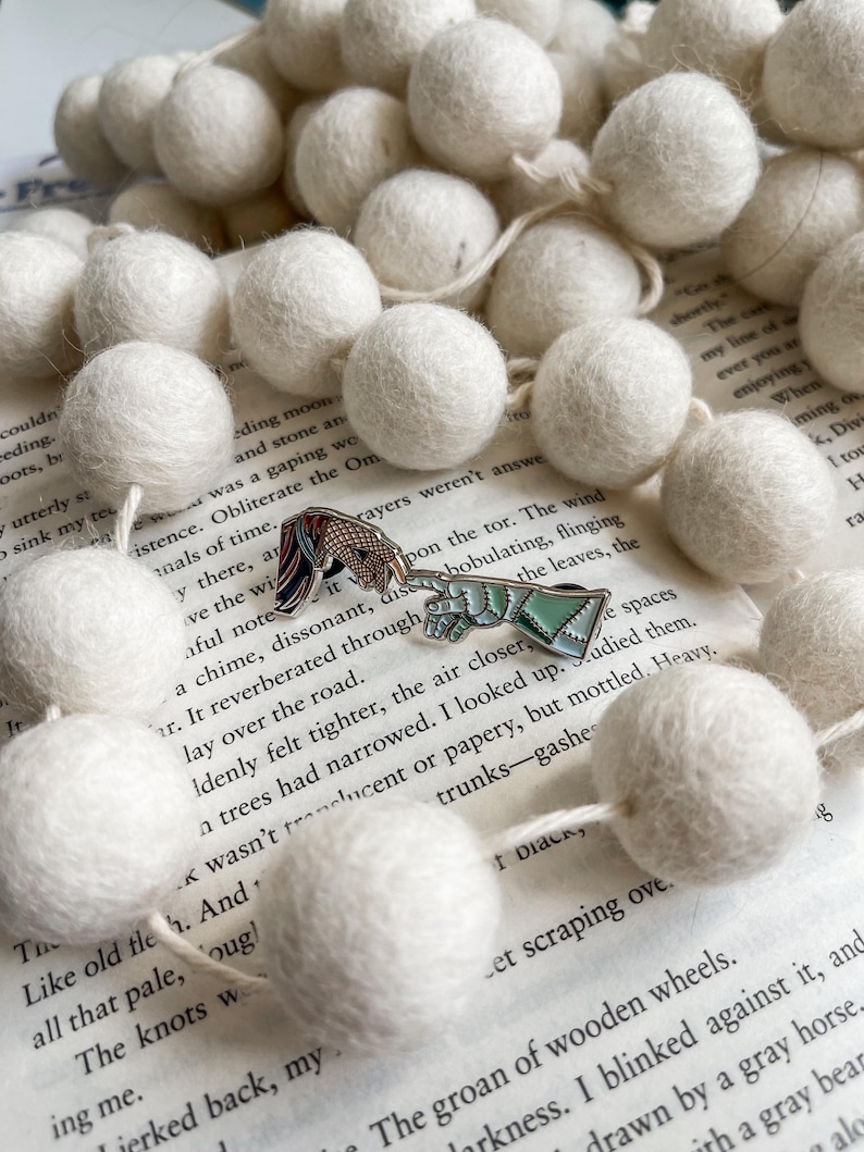 May include: Two enamel pins with detailed designs, one of a horse and the other of a mythical creature, rest on an open book. A garland of white felt balls surrounds the pins. The pins are colorful and the felt balls are soft.