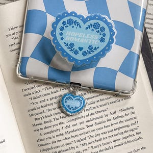 May include: A clear phone case with a blue and white checkered pattern, holding a blue heart-shaped phone grip. The grip has a scalloped edge and the words "HOPELESS ROMANTIC" in the center, surrounded by floral designs. A small heart charm hangs from the grip.