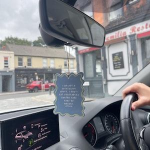 Op de afbeelding: Een lichtblauwe, golvende auto-luchtverfrisser met de tekst "I AM NOT A SUCCESSFUL ADULT. I DON'T EAT VEGETABLES AND/OR TAKE CARE OF MYSELF." hangt aan de achteruitkijkspiegel van een auto. Het dashboard en een deel van het stuurwiel van de auto zijn zichtbaar.