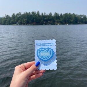 May include: A light blue phone grip with the words "HOPELESS ROMANTIC" and a heart design. The grip is held up against a lake and trees.
