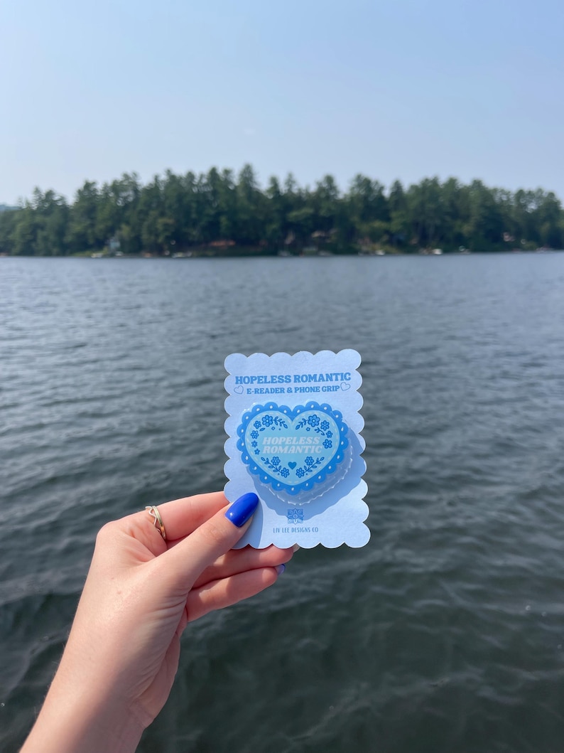 May include: A light blue phone grip with the words "HOPELESS ROMANTIC" and a heart design. The grip is held up against a lake and trees.