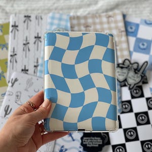 Può includere: Una custodia trasparente per telefono con un motivo a scacchiera ondulato blu e crema. Sullo sfondo sono visibili altre custodie con vari motivi, tra cui cactus, ferri di cavallo e faccine sorridenti.