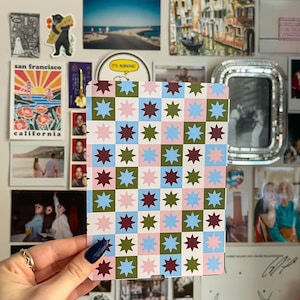 May include: A patterned card featuring a grid of star shapes in pink, blue, green, and burgundy. The card is held against a wall adorned with photographs and stickers, creating a collage-like backdrop.