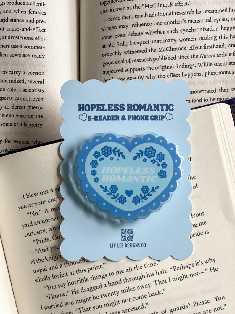 May include: A light blue phone grip and e-reader accessory with a heart-shaped design. The heart features the words "HOPELESS ROMANTIC" and floral accents. The accessory is on a light blue card with the Liv Lee Designs Co. logo.
