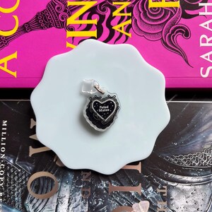 May include: A heart-shaped keychain with the words "Fated Mates" in black script, adorned with a black lace design and silver glitter. The keychain is on a light blue, irregular-shaped plate, with a pink book in the background.