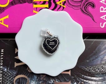 May include: A heart-shaped keychain with the words "Fated Mates" in black script, adorned with a black lace design and silver glitter. The keychain is on a light blue, irregular-shaped plate, with a pink book in the background.