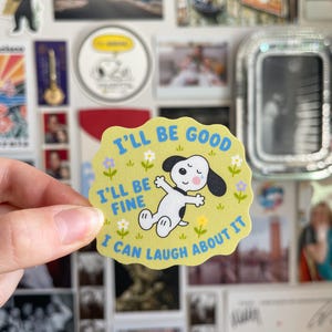May include: A light green, scalloped-edge sticker featuring Snoopy with the text "I'll be good, I'll be fine, I can laugh about it" in blue. The sticker has small flowers and green leaves.