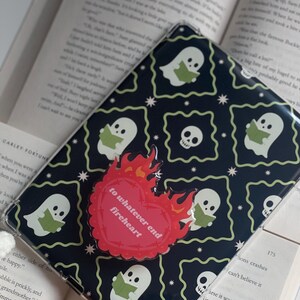 May include: A black phone case featuring a pattern of reading ghosts and skulls. A red heart-shaped sticker with the text "to whatever end fireheart" is attached. The case rests on an open book.