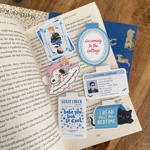 May include: A collection of colorful, whimsical bookmarks and stickers on a wooden surface. The bookmarks feature various designs, including a hockey player, doggy, and phrases like to the cottage and "I read past my bedtime."