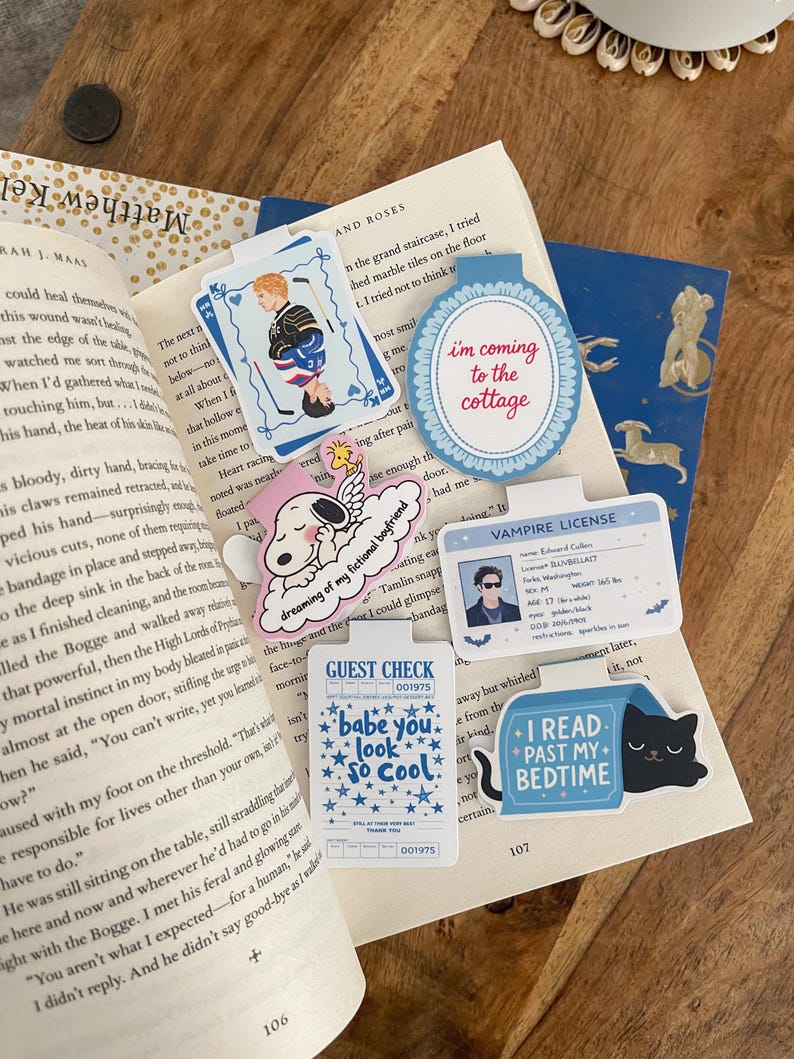May include: A collection of colorful, whimsical bookmarks and stickers on a wooden surface. The bookmarks feature various designs, including a hockey player, doggy, and phrases like to the cottage and "I read past my bedtime."