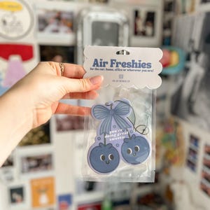 Puede incluir: Ambientador en un paquete de pl&aacute;stico transparente. El ambientador tiene forma de dos ar&aacute;ndanos con caras sonrientes y un lazo. El texto en el embalaje dice "Air Freshies" y "you're doing great sweetie."