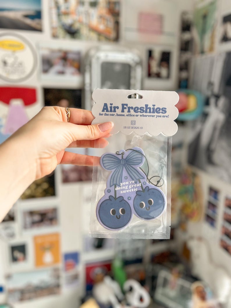Puede incluir: Ambientador en un paquete de pl&aacute;stico transparente. El ambientador tiene forma de dos ar&aacute;ndanos con caras sonrientes y un lazo. El texto en el embalaje dice "Air Freshies" y "you're doing great sweetie."