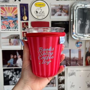 May include: A red and pink striped neoprene cup sleeve featuring the text "Books Spicy Coffee Icy" in light blue script with small yellow star accents. The sleeve has a white brand tag on the side and is fitted around a clear plastic cup.