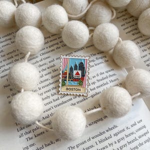 May include: Off-white felt ball garland with a Boston-themed enamel pin. The pin shows a cityscape with "BOSTON" at the base. The garland is arranged on an open book, with text visible.