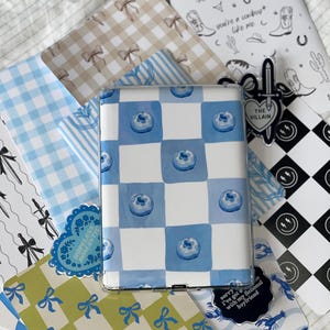Puede incluir: Una funda transparente para tableta con un patrón de cuadros azules y blancos con arándanos en acuarela. Alrededor de la funda hay varios papeles estampados con lazos, rayas e ilustraciones de temática vaquera. También es visible una pegatina con el texto "THE VILLAIN".