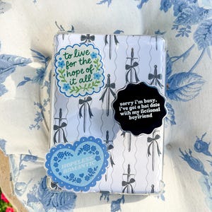 May include: A rectangular clear case featuring a white background with a repeating pattern of black swords tied with bows. Three stickers are attached: one reads to live for the hope of it all, one says sorry i'm busy, i've got a hot date with my fictional boyfriend, and one says HOPELESS ROMANTIC.