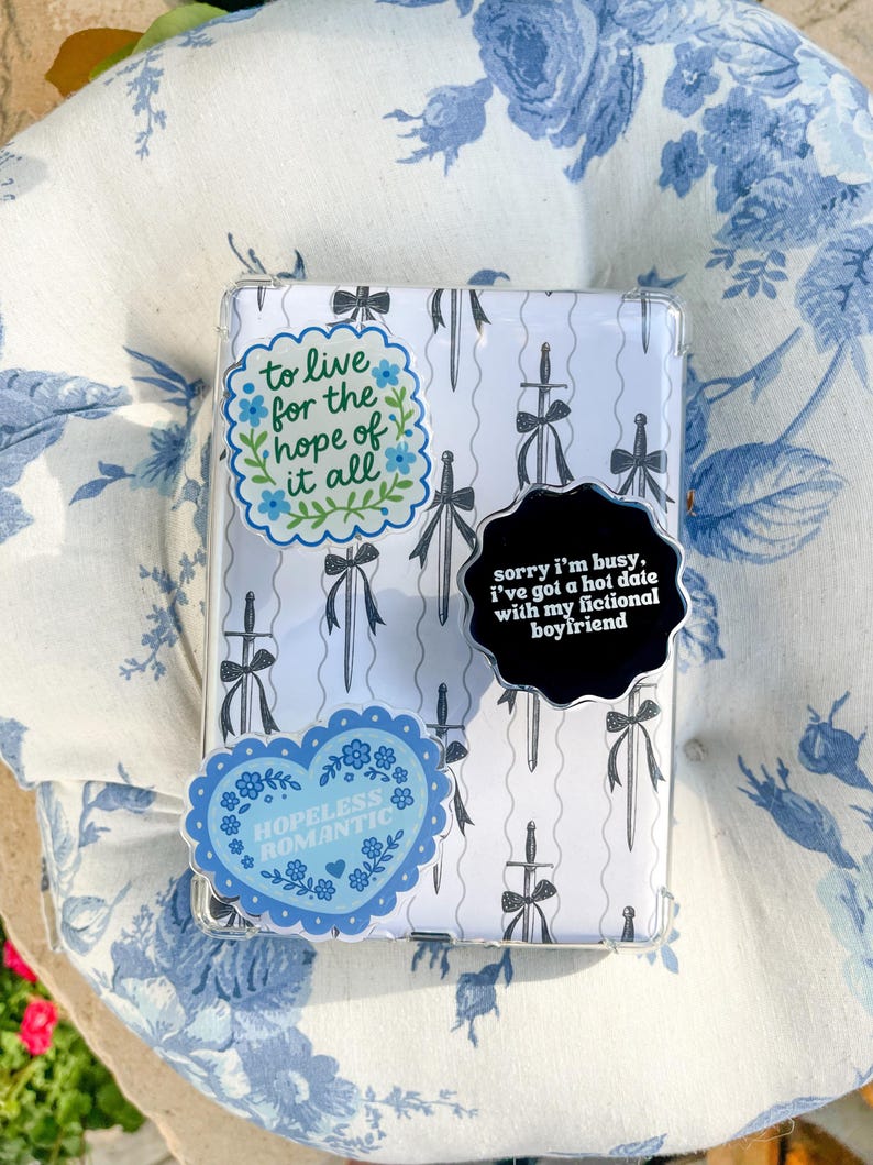 May include: A rectangular clear case featuring a white background with a repeating pattern of black swords tied with bows. Three stickers are attached: one reads to live for the hope of it all, one says sorry i'm busy, i've got a hot date with my fictional boyfriend, and one says HOPELESS ROMANTIC.