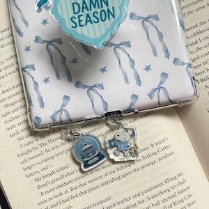 May include: A clear phone case with a blue bow and star pattern, featuring a pop socket with the text "'TIS THE DAMN SEASON". Two clear acrylic charms with winter and cartoon character designs are attached.
