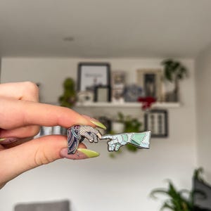 May include: Two enamel pins depicting the iconic 'Creation of Adam' scene. One pin features a hand with a blue and white design, the other a hand with a green and white design. The pins are held up in front of a white wall.