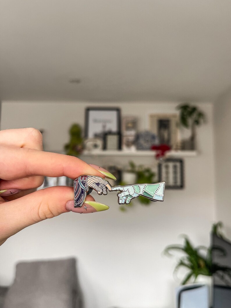May include: Two enamel pins depicting the iconic 'Creation of Adam' scene. One pin features a hand with a blue and white design, the other a hand with a green and white design. The pins are held up in front of a white wall.