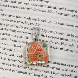 May include: A clear acrylic charm featuring a gingerbread house with the word "Bookstore" on the front. The charm has a festive design with a gingerbread man, Christmas trees, and red and green accents. It hangs from a silver-colored chain.