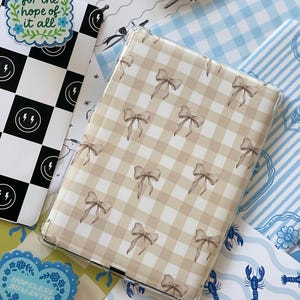 Op de afbeelding: Een doorzichtige tablethoes met een beige en wit geruit patroon en bruine strikken. Rondom de hoes liggen diverse papieren en stickers met patronen, waaronder een zwart-wit dambordpatroon, blauw-witte bloemen en de tekst "to live for the hope of it all."