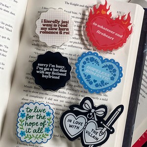 May include: A collection of acrylic pins with various designs and text, including phrases like "I literally just want to read my slow burn romance & rot" and “hopeless romantic”The pins are in different shapes and colors.