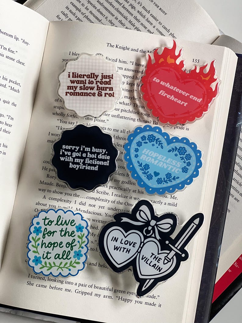 May include: A collection of acrylic pins with various designs and text, including phrases like "I literally just want to read my slow burn romance & rot" and “hopeless romantic”The pins are in different shapes and colors.