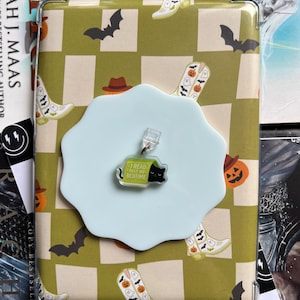 May include: A clear tablet case with a green and white checked pattern, decorated with Halloween motifs. A light blue charm, shaped like a cloud, holds a black cat and the text "I read past my bedtime".