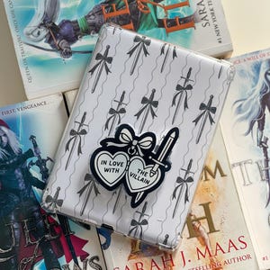May include: A clear phone case with a pattern of swords and bows, and a pop socket that says "In Love With The Villain" is placed on top of several books. The books are by Sarah J. Maas, with titles like "Fire" and "Queen of Shadows".