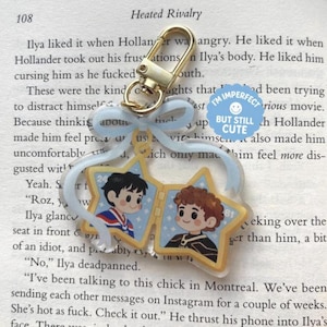 May include: A star-shaped keyring with two cartoon portraits, a gold-coloured clasp, and a light blue ribbon. The keyring has the text "I'm imperfect but still cute" on a blue circle. The background is a book page.