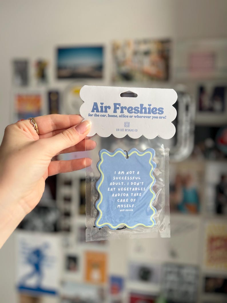 May include: A blue air freshener with a wavy yellow border, packaged in clear plastic. The text reads "I AM NOT A SUCCESSFUL ADULT. I DON'T EAT VEGETABLES AND/OR TAKE CARE OF MYSELF." The packaging says "Air Freshies".
