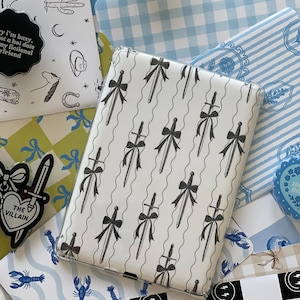 May include: A collection of decorative items with various patterns. A white case with a black sword and bow design is in the center. Other items include a pin with the text "The Villain" and a pin with the text "Sorry I'm busy, not a hot date, my fictional boyfriend."