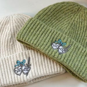 Pode incluir: São exibidas duas toucas de malha canelada, uma creme e outra verde. Cada touca apresenta um patch bordado com dois corações, uma espada e o texto "IN LOVE WITH THE VILLAIN". Um laço azul adorna o patch.