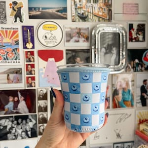 Könnte beinhalten: Eine blau-weiß karierte Becherhülle mit Blaubeer-Designs. Die Hülle besteht aus einem weichen, isolierenden Material. Die Becherhülle ist für eine Tasse in Standardgröße konzipiert.