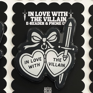 May include: A black and white acrylic accessory featuring a heart-shaped locket design with a bow on top and a sword piercing the right side. The text reads IN LOVE WITH THE VILLAIN. It is mounted on a black scalloped card labeled LIV LEE DESIGNS CO.