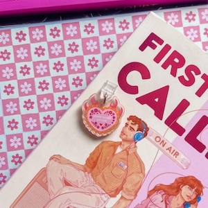 May include: A clear acrylic charm with a heart-shaped design and the text "SLOW BURN LOVER" sits atop a book with the words "FIRST CALL" and an illustration of a man and woman. The background includes a pink floral pattern.