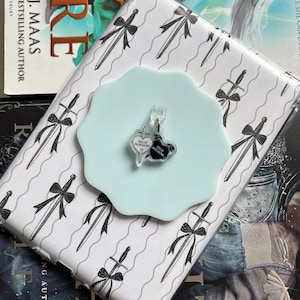 Puede incluir: Una funda transparente para teléfono con un patrón de espadas blancas y negras, un soporte para teléfono de color azul claro y un llavero con el texto "Hello Princeling, Hello Witchling" se muestran encima de un libro. El libro tiene el título "Fire" y el nombre de la autora, Sarah J. Maas.