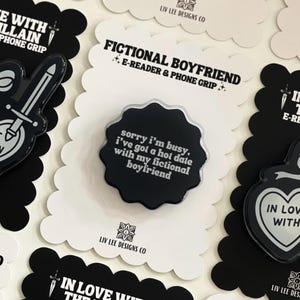 May include: A black scalloped-edge phone grip on a white backing card. The grip features the white text: sorry i'm busy, i've got a hot date with my fictional boyfriend. The backing card displays the text: FICTIONAL BOYFRIEND E-READER & PHONE GRIP.
