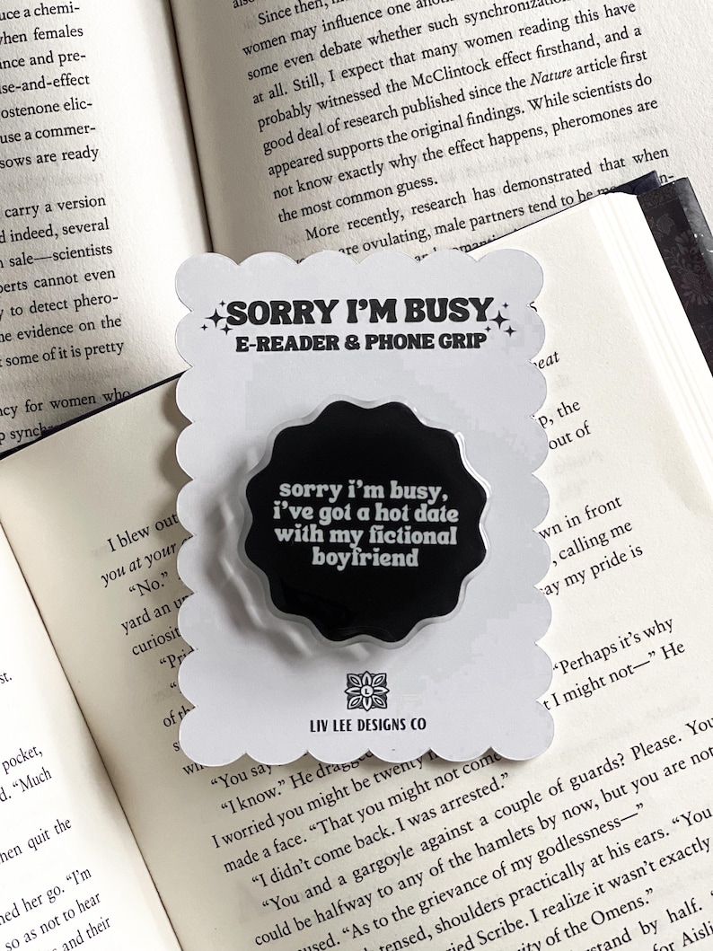 May include: A white, scalloped-edge phone grip with the text "SORRY I'M BUSY E-READER & PHONE GRIP" and a black circle with the text "sorry i'm busy, i've got a hot date with my fictional boyfriend." The grip is on an open book.