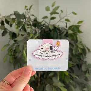 May include: A magnetic bookmark featuring a puppy sleeping on a cloud with the text "dreaming of my fictional boyfriend."The bookmark has a pink border and the words "MAGNETIC BOOKMARK" at the bottom.