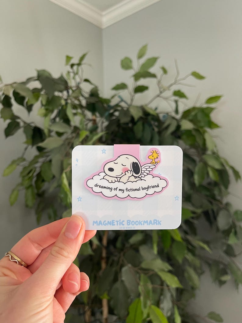 May include: A magnetic bookmark featuring a puppy sleeping on a cloud with the text "dreaming of my fictional boyfriend."The bookmark has a pink border and the words "MAGNETIC BOOKMARK" at the bottom.