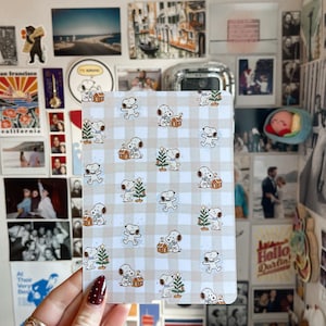 Peut inclure: Un bloc-notes rectangulaire avec un motif répété de personnages Snoopy avec des arbres de Noël et des maisons sur un fond à carreaux beige et blanc. Le bloc-notes est tenu dans une main avec du vernis à ongles bordeaux.